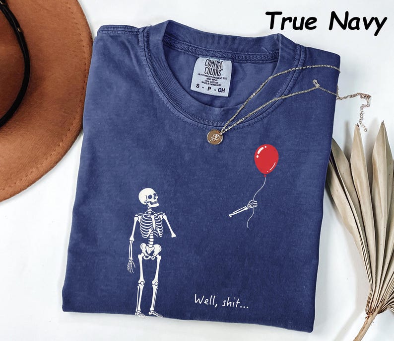 May include: A true navy blue t-shirt featuring a white graphic of a skeleton looking at a detached skeletal hand holding a red balloon. The text "Well, shit..." is printed in white below the graphic. The shirt has a Comfort Colors brand label.