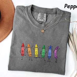 May include: A pepper-colored t-shirt featuring a graphic of six anthropomorphic crayons in red, orange, yellow, green, blue, and violet. Each crayon has a smiling face and stick-figure limbs. The shirt has a Comfort Colors brand label.