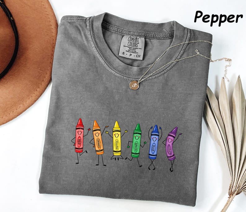 May include: A pepper-colored t-shirt featuring a graphic of six anthropomorphic crayons in red, orange, yellow, green, blue, and violet. Each crayon has a smiling face and stick-figure limbs. The shirt has a Comfort Colors brand label.