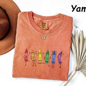 May include: A yam-colored t-shirt featuring a graphic of six anthropomorphic crayons in red, orange, yellow, green, blue, and purple, each with a smiling face and limbs. The shirt has a Comfort Colors brand label at the inner neckline.