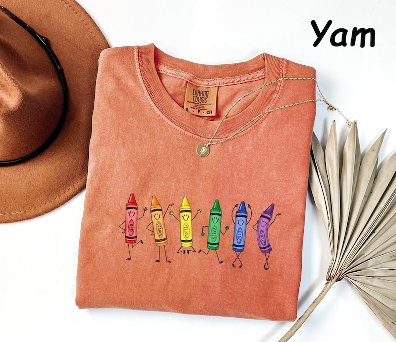 May include: A yam-colored t-shirt featuring a graphic of six anthropomorphic crayons in red, orange, yellow, green, blue, and purple, each with a smiling face and limbs. The shirt has a Comfort Colors brand label at the inner neckline.