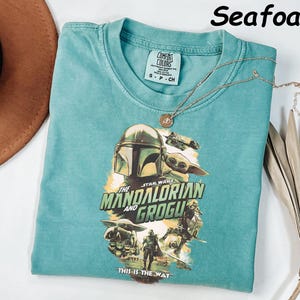 May include: Seafoam green t-shirt featuring a graphic of The Mandalorian and Grogu from Star Wars. The shirt has a crew neck and short sleeves. A gold necklace with a pendant rests on the shirt. The text "THIS IS THE WAY" is printed on the shirt.