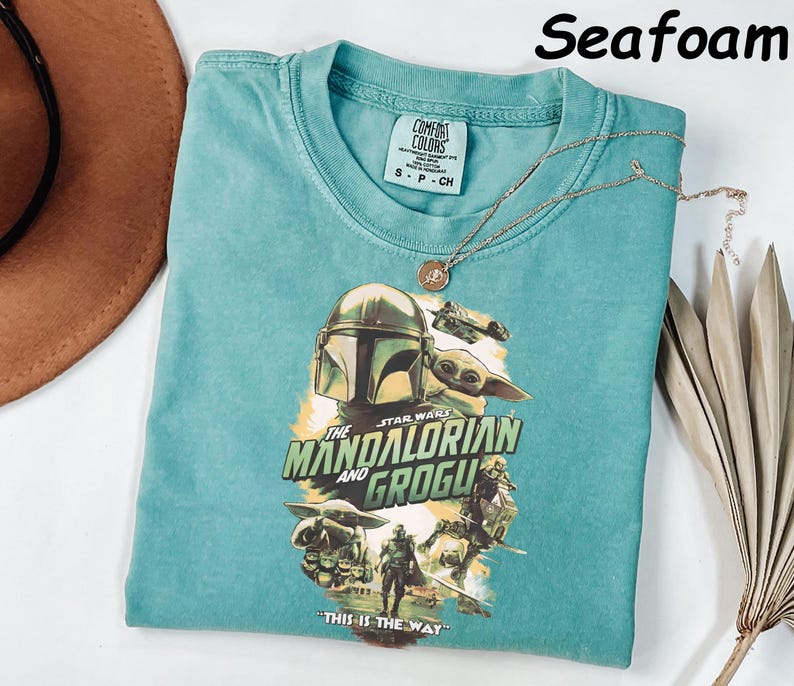 May include: Seafoam green t-shirt featuring a graphic of The Mandalorian and Grogu from Star Wars. The shirt has a crew neck and short sleeves. A gold necklace with a pendant rests on the shirt. The text "THIS IS THE WAY" is printed on the shirt.