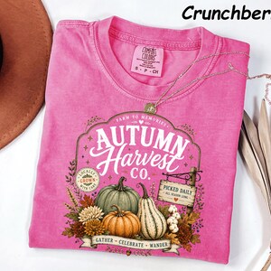 May include: A bright pink short-sleeve t-shirt featuring a graphic print with the text Autumn Harvest Co., Farm to Memories, Locally Grown, and Picked Daily. The design includes illustrations of pumpkins, gourds, and autumn foliage.