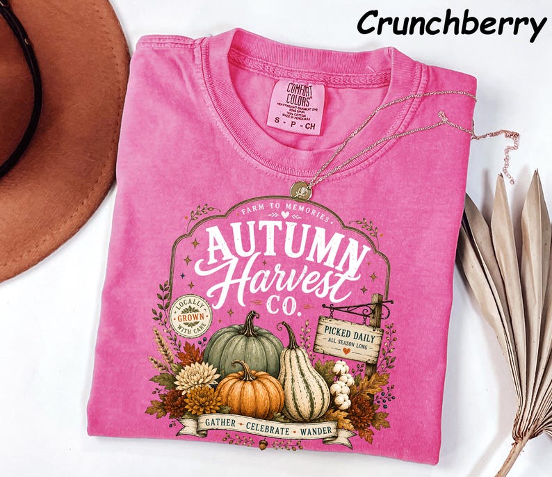 May include: A bright pink short-sleeve t-shirt featuring a graphic print with the text Autumn Harvest Co., Farm to Memories, Locally Grown, and Picked Daily. The design includes illustrations of pumpkins, gourds, and autumn foliage.