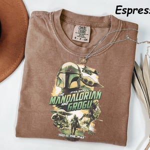May include: Espresso-colored t-shirt featuring a graphic of The Mandalorian and Grogu from Star Wars. The shirt has a crew neck and short sleeves. A silver necklace with a pendant rests on the shirt. The text "THIS IS THE WAY" is visible.