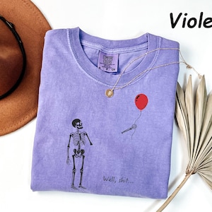May include: A violet short-sleeved t-shirt featuring a graphic of a skeleton looking at a red balloon held by a detached skeletal hand. The text "Well, shit..." is printed below the graphic. The shirt has a Comfort Colors brand label.