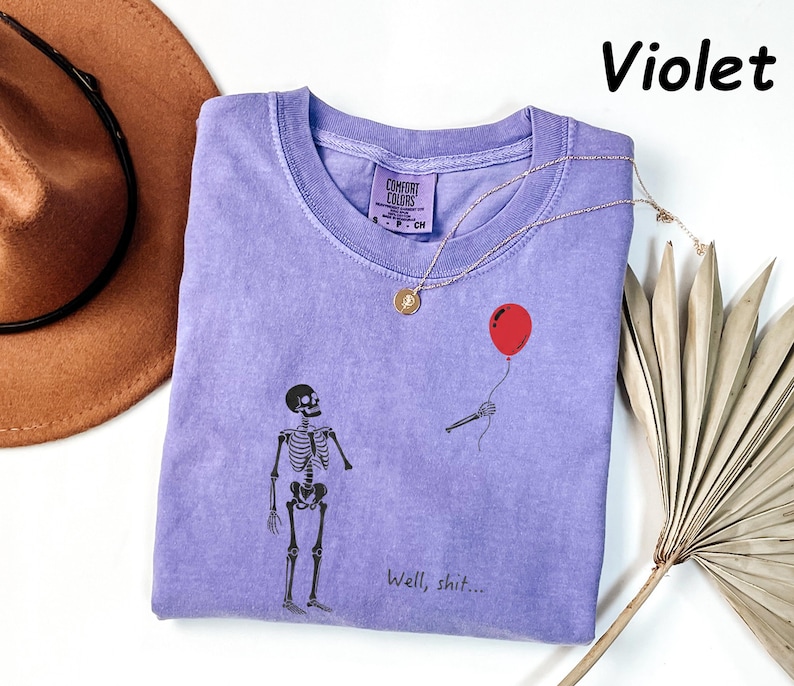 May include: A violet short-sleeved t-shirt featuring a graphic of a skeleton looking at a red balloon held by a detached skeletal hand. The text "Well, shit..." is printed below the graphic. The shirt has a Comfort Colors brand label.