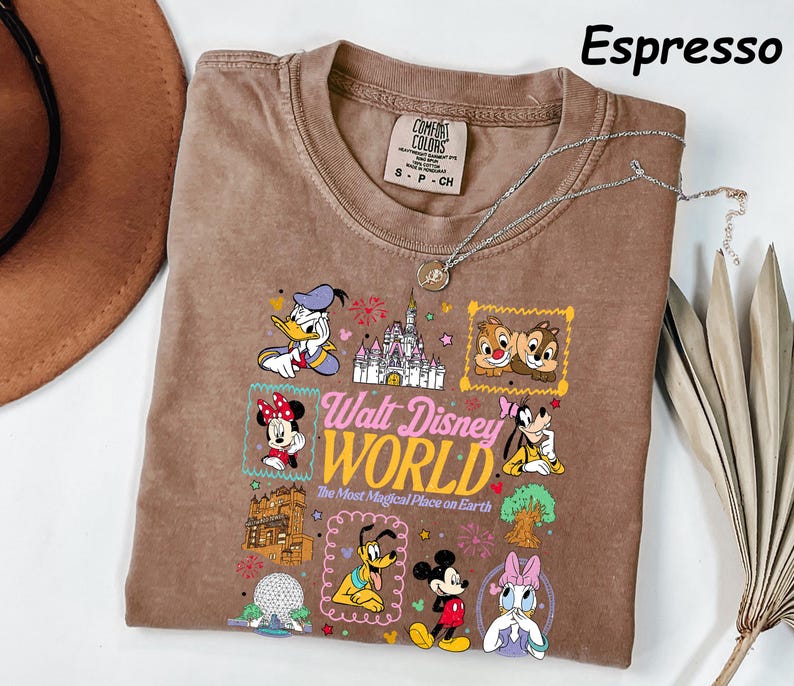 K&ouml;nnte beinhalten: Espressofarbenes T-Shirt mit einem farbenfrohen Disney World-Grafikdruck, darunter Donald Duck und Mickey Mouse. Der Text "Walt Disney World" ist zu sehen. Eine silberne Halskette liegt auf dem Shirt. Gr&ouml;&szlig;e S-P-CH.