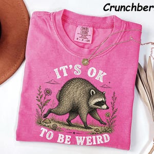 May include: A bright pink short-sleeve t-shirt featuring a graphic of a raccoon walking among flowers and mushrooms. The text reads IT'S OK TO BE WEIRD. The shirt has a Comfort Colors label at the neck, and the colour is labelled as Crunchberry.
