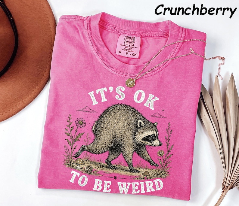 May include: A bright pink short-sleeve t-shirt featuring a graphic of a raccoon walking among flowers and mushrooms. The text reads IT'S OK TO BE WEIRD. The shirt has a Comfort Colors label at the neck, and the colour is labelled as Crunchberry.