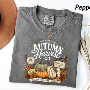 May include: A pepper-colored t-shirt featuring a graphic with the text Autumn Harvest Co., Farm to Memories, Locally Grown With Care, Picked Daily All Season Long, and Gather Celebrate Wander, along with illustrations of pumpkins and autumn foliage.