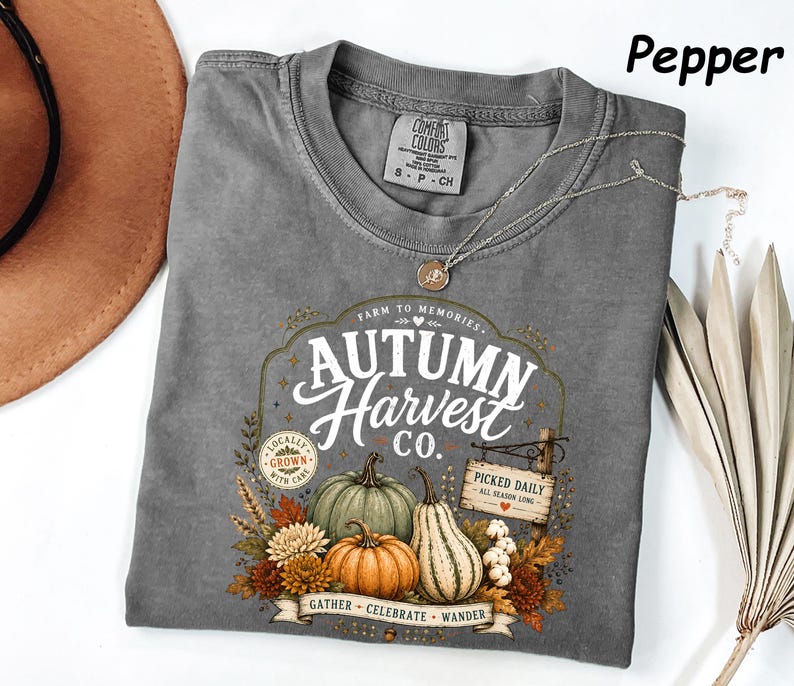 May include: A pepper-colored t-shirt featuring a graphic with the text Autumn Harvest Co., Farm to Memories, Locally Grown With Care, Picked Daily All Season Long, and Gather Celebrate Wander, along with illustrations of pumpkins and autumn foliage.