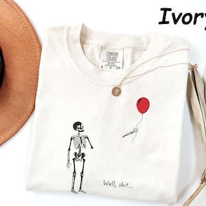 May include: An ivory t-shirt featuring a black graphic of a skeleton standing near a disembodied skeletal hand holding a red balloon. The text "Well, shit..." is printed in black near the bottom. The shirt has a Comfort Colors brand label.