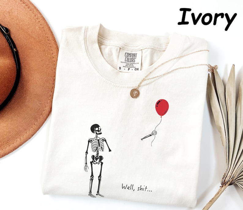 May include: An ivory t-shirt featuring a black graphic of a skeleton standing near a disembodied skeletal hand holding a red balloon. The text "Well, shit..." is printed in black near the bottom. The shirt has a Comfort Colors brand label.