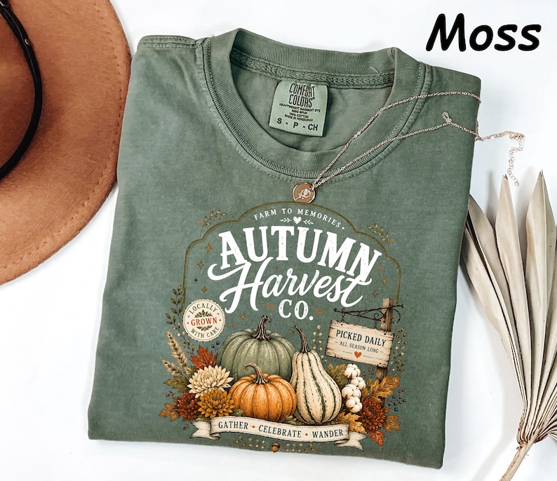 May include: A moss green short-sleeve t-shirt featuring a graphic print with the text Autumn Harvest Co. Farm to Memories. The design includes illustrations of pumpkins, gourds, and autumn flowers, along with a sign reading Picked Daily.