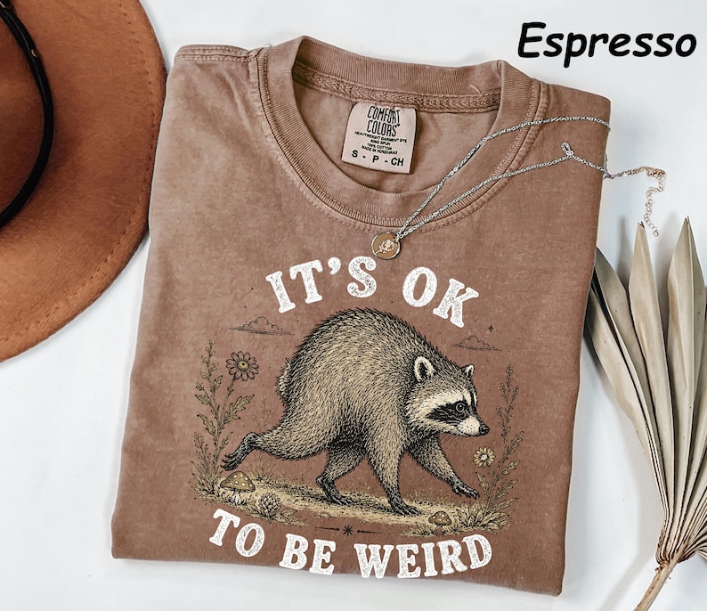May include: An espresso-coloured t-shirt featuring a graphic of a raccoon walking through a field with flowers and mushrooms. The text "IT'S OK TO BE WEIRD" is printed in white above and below the raccoon. The shirt has a Comfort Colors label.