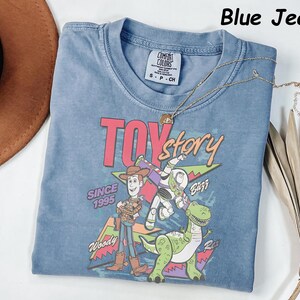 May include: A blue jean colored t-shirt featuring a graphic of Woody, Buzz Lightyear, and Rex from the movie Toy Story. The text "TOY story" is in pink and green, with "SINCE 1995" below. A gold necklace is draped across the shirt.