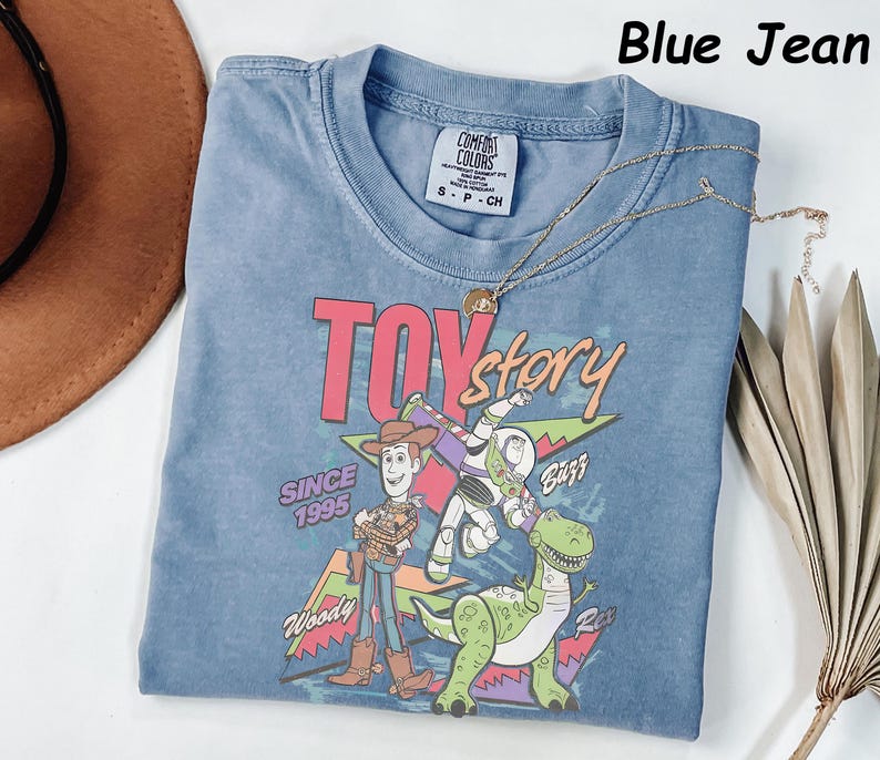 May include: A blue jean colored t-shirt featuring a graphic of Woody, Buzz Lightyear, and Rex from the movie Toy Story. The text "TOY story" is in pink and green, with "SINCE 1995" below. A gold necklace is draped across the shirt.