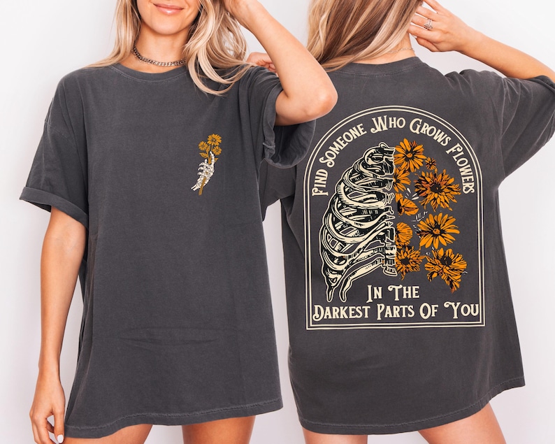 May include: Dark gray t-shirt with a skeleton hand holding yellow flowers on the front. The back of the shirt features a rib cage design with yellow flowers and the text "Find someone who grows flowers in the darkest parts of you."