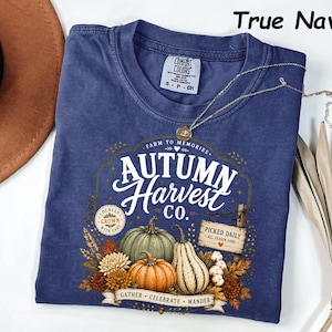 May include: A true navy short-sleeve t-shirt featuring a graphic print with the text Autumn Harvest Co. The design includes illustrations of pumpkins, gourds, autumn flowers, and a wooden sign that reads Picked Daily All Season Long.