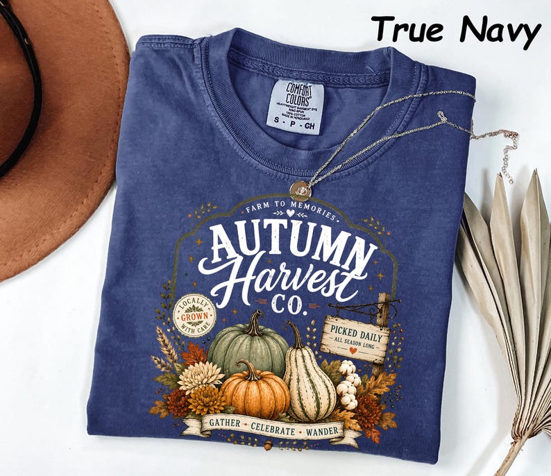 May include: A true navy short-sleeve t-shirt featuring a graphic print with the text Autumn Harvest Co. The design includes illustrations of pumpkins, gourds, autumn flowers, and a wooden sign that reads Picked Daily All Season Long.