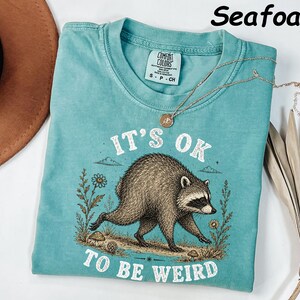 May include: A seafoam green short-sleeve t-shirt featuring a graphic of a raccoon walking through flowers and mushrooms. The text "IT'S OK TO BE WEIRD" is printed in white above and below the raccoon. The shirt has a crew neckline and a tag.