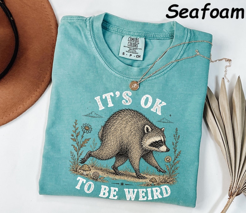 May include: A seafoam green short-sleeve t-shirt featuring a graphic of a raccoon walking through flowers and mushrooms. The text "IT'S OK TO BE WEIRD" is printed in white above and below the raccoon. The shirt has a crew neckline and a tag.