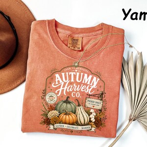 May include: A yam-colored t-shirt featuring a graphic print with the text Autumn Harvest Co., Farm to Memories, Locally Grown, and Picked Daily. The design includes illustrations of pumpkins, gourds, and autumn foliage in a rustic, vintage-inspired style.