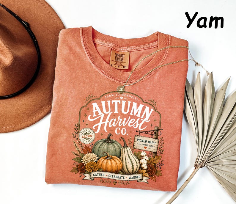 May include: A yam-colored t-shirt featuring a graphic print with the text Autumn Harvest Co., Farm to Memories, Locally Grown, and Picked Daily. The design includes illustrations of pumpkins, gourds, and autumn foliage in a rustic, vintage-inspired style.