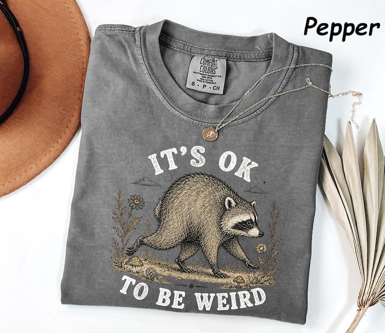 May include: A pepper-coloured t-shirt featuring a graphic of a raccoon walking through a field with flowers and mushrooms. The text above and below the raccoon reads, IT'S OK TO BE WEIRD. The shirt has a crew neckline and a Comfort Colors brand label.