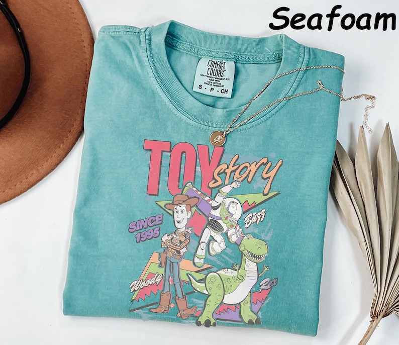 May include: Seafoam green t-shirt featuring a graphic of Woody, Buzz Lightyear, and Rex from Toy Story. The shirt has the text "TOY STORY" and "SINCE 1995". A gold necklace with a pendant is on the shirt.