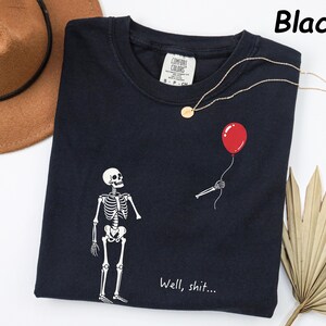 May include: A black t-shirt featuring a white skeleton graphic, a detached skeletal hand holding a red balloon, and the text "Well, shit..." printed in white. The shirt has a Comfort Colors brand label at the inner neckline and a crew neck collar.