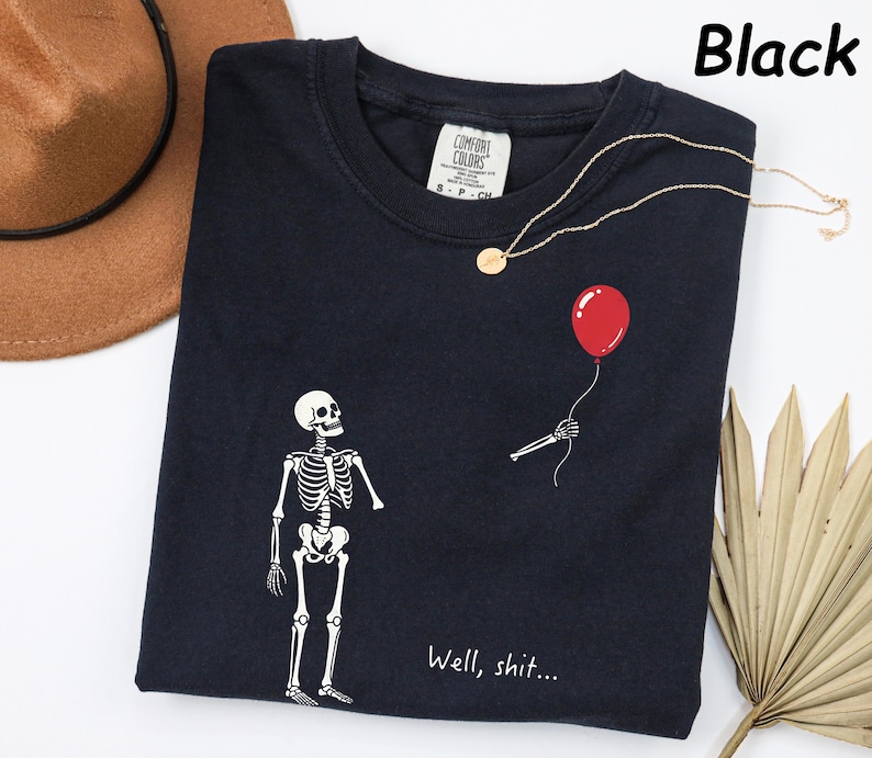 May include: A black t-shirt featuring a white skeleton graphic, a detached skeletal hand holding a red balloon, and the text "Well, shit..." printed in white. The shirt has a Comfort Colors brand label at the inner neckline and a crew neck collar.
