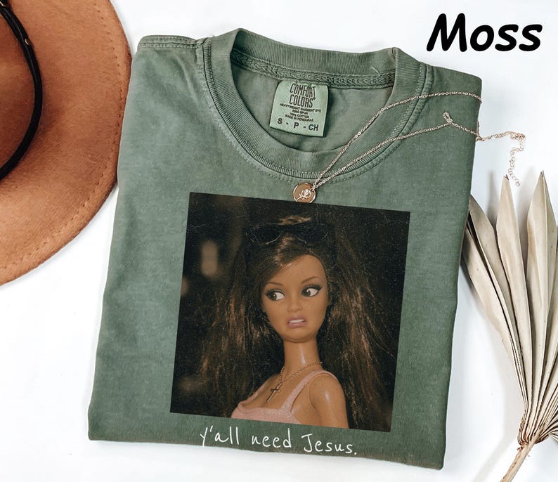 May include: A moss green crew neck t-shirt featuring a graphic print of a doll with long brown hair wearing sunglasses and a pink top. The text Y'all need Jesus is printed in white below the graphic. The shirt has a Comfort Colors brand label.