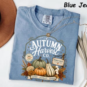 May include: A blue jean colored t-shirt featuring a graphic print with the text Autumn Harvest Co. The design includes illustrations of pumpkins, gourds, autumn leaves, and a wooden sign that reads Picked Daily All Season Long. The shirt has a crew neckline and a Comfort Colors brand label at the inner collar.