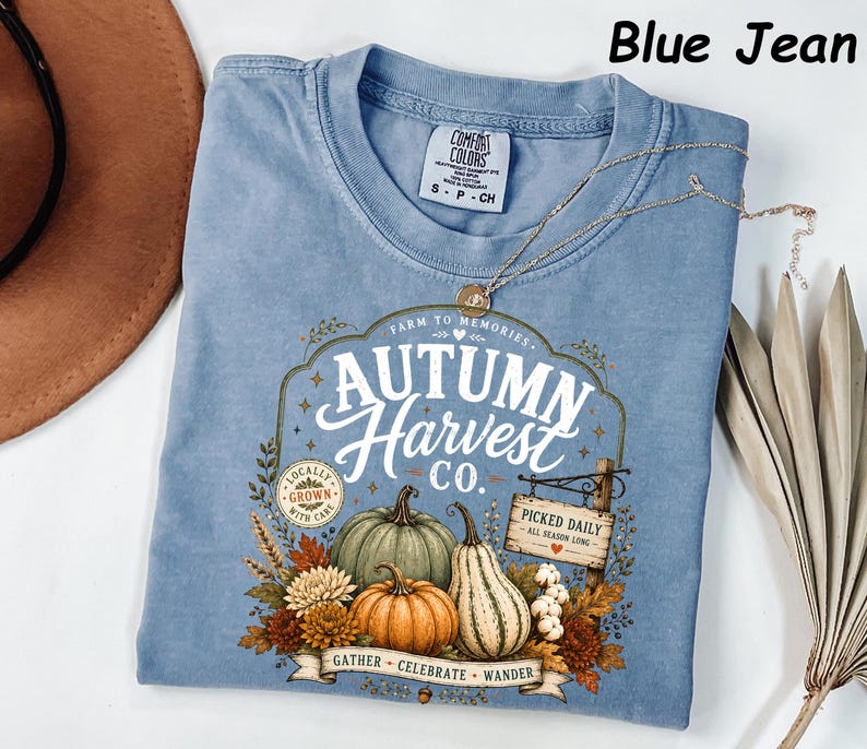 May include: A blue jean colored t-shirt featuring a graphic print with the text Autumn Harvest Co. The design includes illustrations of pumpkins, gourds, autumn leaves, and a wooden sign that reads Picked Daily All Season Long. The shirt has a crew neckline and a Comfort Colors brand label at the inner collar.