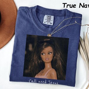 May include: A true navy blue t-shirt featuring a graphic print of a doll with long brown hair wearing sunglasses on her head and a cross necklace. The text "y'all need Jesus." is printed below the graphic. The shirt has a Comfort Colors label.