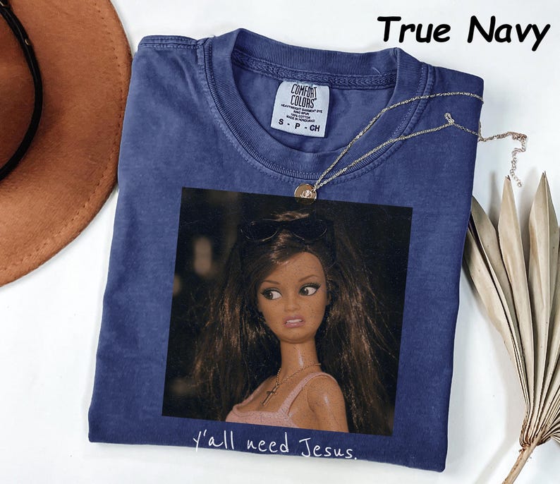 May include: A true navy blue t-shirt featuring a graphic print of a doll with long brown hair wearing sunglasses on her head and a cross necklace. The text "y'all need Jesus." is printed below the graphic. The shirt has a Comfort Colors label.