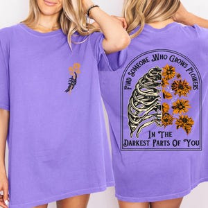May include: A lilac-colored t-shirt with a graphic design. The front features a skeletal hand holding flowers. The back displays a rib cage with flowers and the text "FIND SOMEONE WHO GROWS FLOWERS IN THE DARKEST PARTS OF YOU."
