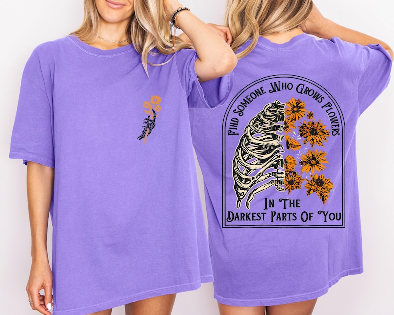 May include: A lilac-colored t-shirt with a graphic design. The front features a skeletal hand holding flowers. The back displays a rib cage with flowers and the text "FIND SOMEONE WHO GROWS FLOWERS IN THE DARKEST PARTS OF YOU."