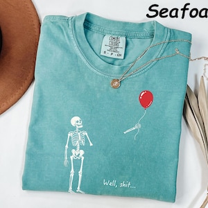 May include: A seafoam green t-shirt featuring a white graphic of a skeleton standing near a detached skeletal arm holding a red balloon. The text "Well, shit..." is printed in white below the graphic. The shirt has a Comfort Colors brand label.
