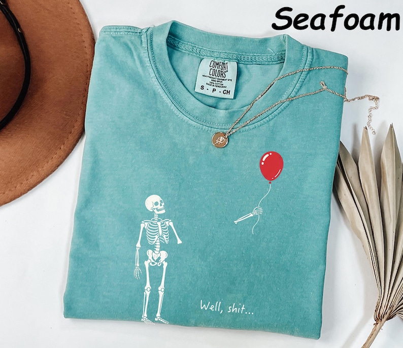 May include: A seafoam green t-shirt featuring a white graphic of a skeleton standing near a detached skeletal arm holding a red balloon. The text "Well, shit..." is printed in white below the graphic. The shirt has a Comfort Colors brand label.