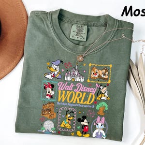 K&ouml;nnte beinhalten: Moosgr&uuml;nes T-Shirt mit einer bunten Grafik von Disney-Figuren und dem Text "Walt Disney World, The Most Magical Place on Earth." Das Shirt hat einen Rundhalsausschnitt und kurze &Auml;rmel. Eine silberne Halskette ist &uuml;ber dem Shirt drapiert.