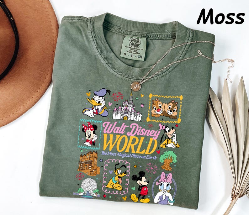 K&ouml;nnte beinhalten: Moosgr&uuml;nes T-Shirt mit einer bunten Grafik von Disney-Figuren und dem Text "Walt Disney World, The Most Magical Place on Earth." Das Shirt hat einen Rundhalsausschnitt und kurze &Auml;rmel. Eine silberne Halskette ist &uuml;ber dem Shirt drapiert.