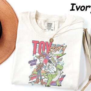 May include: Ivory-colored t-shirt featuring a graphic of Toy Story characters, including Woody, Buzz Lightyear, and Rex. The design includes the text "TOY STORY" and "SINCE 1995". A gold necklace and a brown hat are also visible.