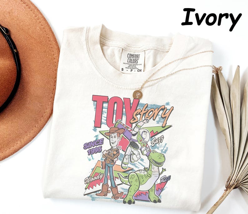 May include: Ivory-colored t-shirt featuring a graphic of Toy Story characters, including Woody, Buzz Lightyear, and Rex. The design includes the text "TOY STORY" and "SINCE 1995". A gold necklace and a brown hat are also visible.