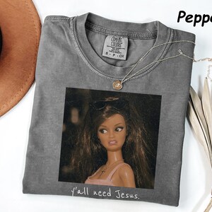 May include: A pepper-colored short-sleeve t-shirt featuring a graphic print of a doll with long brown hair and sunglasses on her head. The doll wears a pink top and a necklace. Below the image, the text reads, y'all need Jesus.