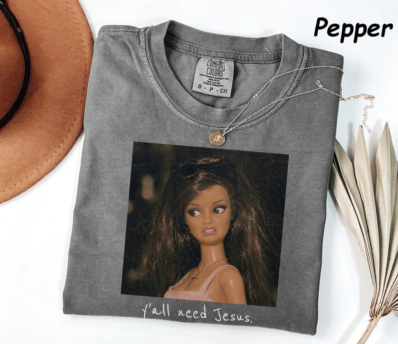 May include: A pepper-colored short-sleeve t-shirt featuring a graphic print of a doll with long brown hair and sunglasses on her head. The doll wears a pink top and a necklace. Below the image, the text reads, y'all need Jesus.
