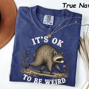 May include: A true navy blue short-sleeve t-shirt featuring a graphic of a raccoon walking through a field with mushrooms and flowers. The text "IT'S OK TO BE WEIRD" is printed in white above and below the raccoon. The shirt has a Comfort Colors label.